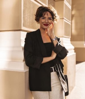 Joyful lady in jacket and white pants smiling outside. Lovely woman with bright lips in eyeglasses looking into camera outdoors.